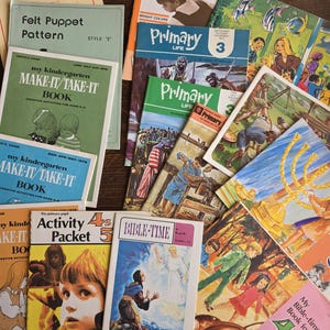 May include: A collection of vintage children's books and activity packets. Titles include "Primary Life," "Bible-Time," and "Make-It/Take-It Book." The covers feature colorful illustrations and text, with a variety of sizes and shapes. The books are arranged on a wooden surface.