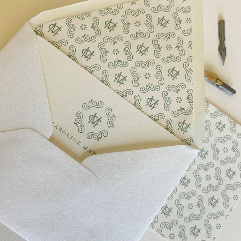 May include: Cream-colored stationery set featuring a patterned design in a muted green. The set includes an envelope, a card with the name "CAROLINE MAT" printed on it, and a patterned card. A calligraphy pen is also visible.
