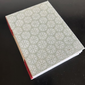 May include: A small, rectangular notebook with a floral pattern cover. The cover is a light green color with a repeating white flower design. The spine is red, and the pages are white.