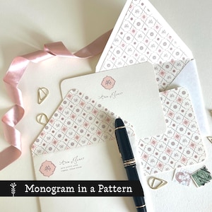 May include: Stationery set featuring a monogram in a pattern. The set includes a card, envelope, and pen. The envelope and card have a pink and white patterned design. The text "Aria Klineo" is visible on the card. Gold paper clips and a pink ribbon are also present.