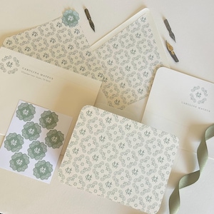 May include: Stationery set featuring cream-colored envelopes, notecards, and stickers with a repeating pattern in a muted green. The set includes a card with the name "CAROLINA MATEUS" printed on it, along with calligraphy pens and a green ribbon.