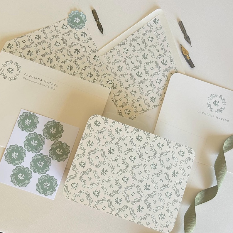 May include: Stationery set featuring cream-colored envelopes, notecards, and stickers with a repeating pattern in a muted green. The set includes a card with the name "CAROLINA MATEUS" printed on it, along with calligraphy pens and a green ribbon.