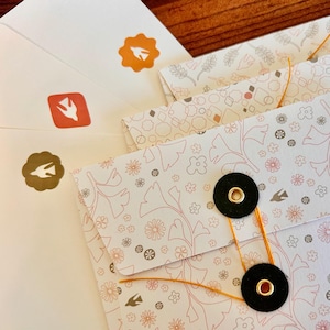 May include: Close-up of stationery items, including envelopes and patterned paper. The envelopes are white with orange and brown stickers featuring a bird design. The patterned paper has floral and geometric designs in shades of pink, gray, and brown. The paper is secured with black buttons and orange string.