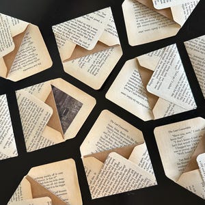May include: Overhead shot of several small, handmade paper envelopes made from aged book pages. Some envelopes are open, revealing a glimpse of a photograph. The envelopes are scattered on a dark surface.