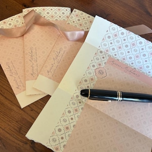 May include: Stationery set featuring pale pink envelopes and notecards with a repeating geometric pattern. A black fountain pen with gold accents rests on the stationery. The envelopes have a ribbon closure and the name "Monica Michliano" printed on them.