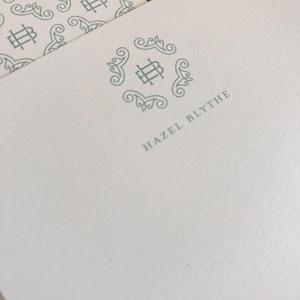 May include: Off-white stationery with a repeating pattern and a centered logo. The logo features the initials "HB" within a decorative circular frame. The name "HAZEL BLYTHE" is printed below the logo in a matching gray color.