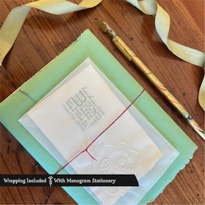 May include: Stationery set with a light green envelope, a white vellum paper, and a red string. A vintage-style pen rests on the wooden surface. The vellum paper has the text "Bound Bliss" and "created by Chrissy". A light green ribbon is also included.