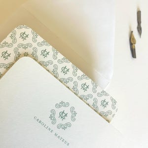 May include: Stationery set featuring a cream-colored card with the name "CAROLINE MATEUS" and a decorative teal monogram. The set includes a matching envelope and a patterned liner. Two vintage pen nibs are also visible.