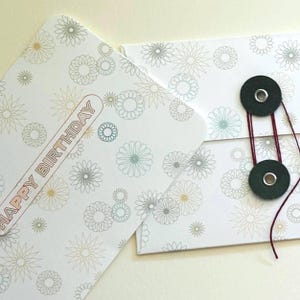 May include: White birthday card with a floral pattern in muted colors. The card features the text "HAPPY BIRTHDAY" in a brown font. The card is secured with two dark green buttons and a red string.