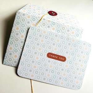 May include: A stationery set featuring a patterned envelope and a card with the words "THANK YOU". The envelope has a button closure and a yellow string. The card and envelope have a repeating floral design in shades of blue, yellow, and white.
