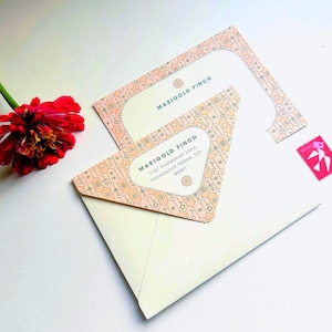 May include: An ivory envelope with a peach and gray patterned design, featuring the text "MARIGOLD FINCH" and address details. A red flower and a pink stamp are also visible.