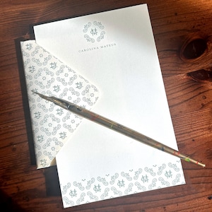 May include: Stationery set featuring a white letter sheet and envelope with a repeating teal pattern and the name "CAROLINA MATEUS". A pen with a colorful design rests on the stationery, all on a wooden surface.