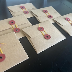 May include: Kraft paper envelopes with a flap closure. Each envelope is secured with two red circular fasteners connected by a yellow string. The envelopes are arranged on a dark surface.