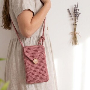 May include: A dusty rose crochet crossbody phone bag with a flap closure and a wooden button. The bag has a long, braided strap. A bundle of dried lavender is tied with twine on a white wall.