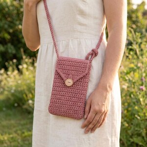 May include: A dusty rose crochet phone bag with a wooden button closure. The bag has a long strap for crossbody wear. The bag is approximately 7 inches tall and 4 inches wide.