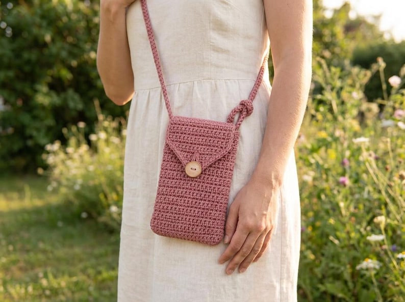 May include: A dusty rose crochet phone bag with a wooden button closure. The bag has a long strap for crossbody wear. The bag is approximately 7 inches tall and 4 inches wide.