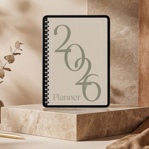 Puede incluir: Una agenda con espiral con el año "2026" en verde salvia y la palabra "Planner" debajo. La agenda se muestra sobre un bloque de mármol, con un jarrón de hojas secas y un bolígrafo dorado cerca. El fondo presenta una paleta de colores neutros.