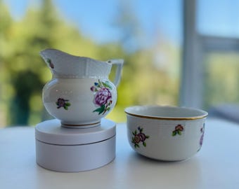 May include: A white porcelain creamer and bowl set. The creamer has a handle and spout, and both pieces are decorated with purple and pink floral designs and gold trim. The set is on a white surface.