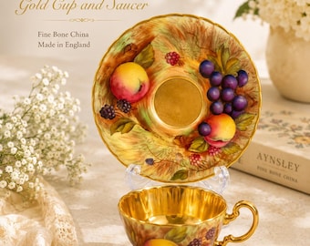 Può includere: Un set di tazza e piattino in fine bone china di Aynsley, modello Orchard Gold. Il piattino presenta un motivo a frutta dipinta con uva e pesche, mentre la tazza ha un interno dorato e un design a frutta coordinato.