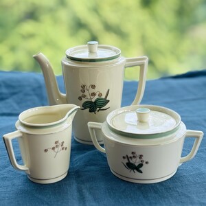 May include: A vintage cream-coloured tea set comprising a teapot, creamer jug, and sugar bowl. Each piece features a green trim and a floral pattern with green leaves and brown berries. The set is on a blue surface.