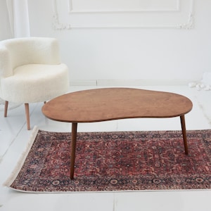 May include: A mid-century modern style living room scene featuring a kidney-shaped wooden coffee table with dark brown legs, set on a patterned rug. A white, fluffy armchair and a fireplace are in the background.