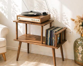 May include: A two-tiered wooden record player stand with tapered legs, featuring a dark wood finish. The top shelf holds a record player with a clear dust cover, while the bottom shelf contains a collection of vinyl records in a side rack.