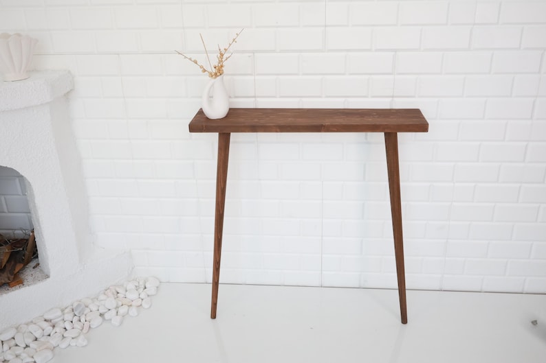 May include: A mid-century modern style console table with a dark brown wooden top and legs. The table is 48 inches long and 12 inches wide. A white vase with dried flowers sits on the table. The table is against a white brick wall.
