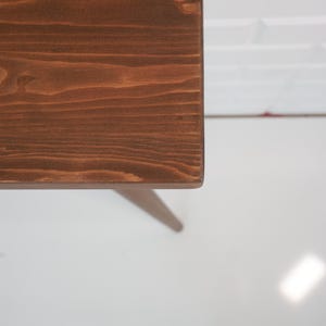 May include: A close-up of a wooden table corner. The table has a rich, dark brown stain, showcasing the natural wood grain. The table leg is visible, and the background is a white wall and floor.