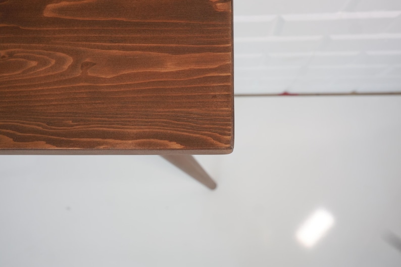 May include: A close-up of a wooden table corner. The table has a rich, dark brown stain, showcasing the natural wood grain. The table leg is visible, and the background is a white wall and floor.