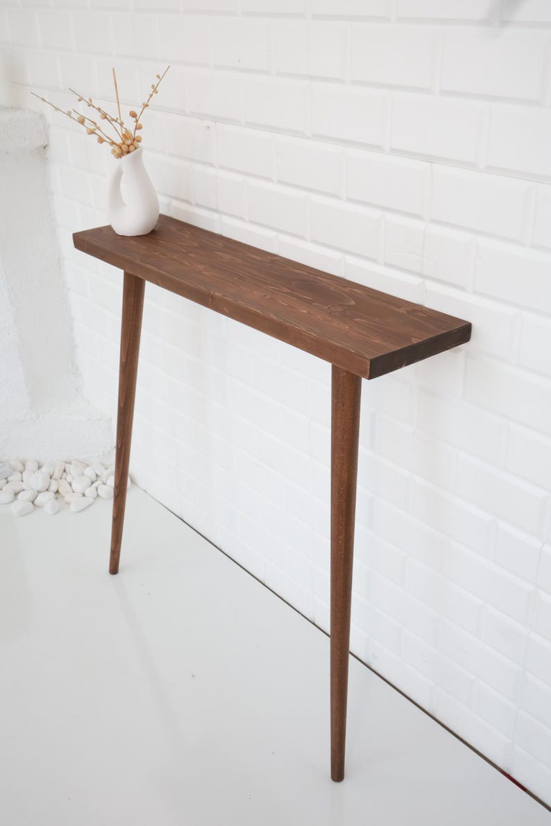 May include: A narrow, dark brown wooden console table with four tapered legs. A small white vase with dried floral stems sits on the tabletop. The table is against a white brick wall.