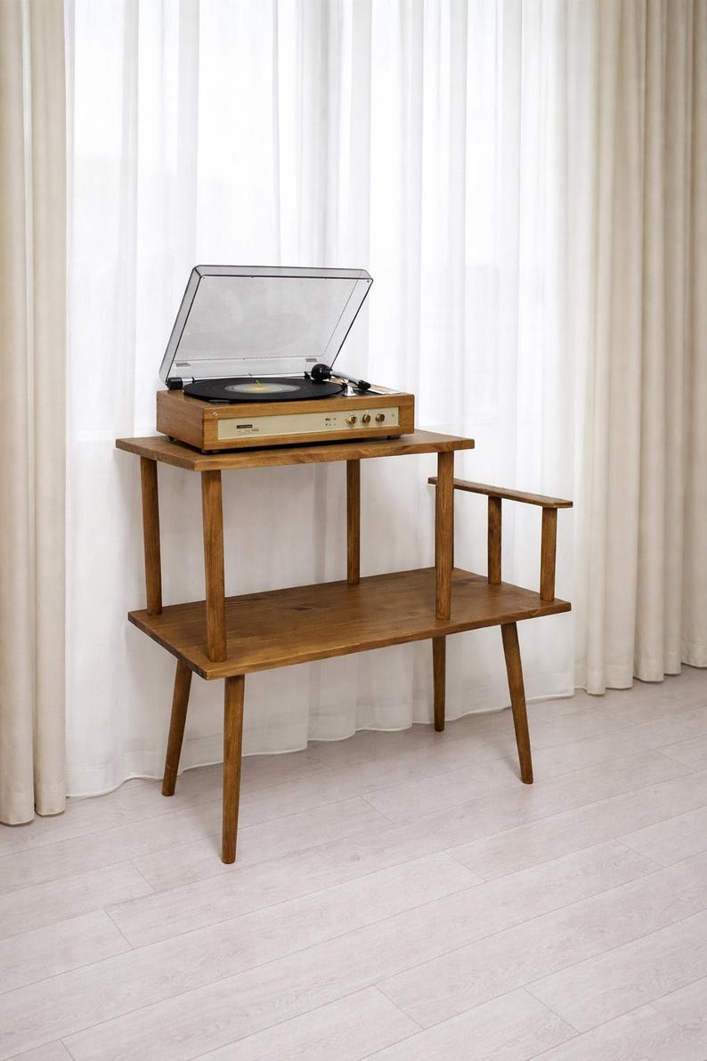 May include: A wooden two-tier stand with a light brown finish, featuring four tapered legs and a small side armrest. A rectangular record player with a clear plastic dust cover and a black turntable platter sits on the top shelf of the stand.