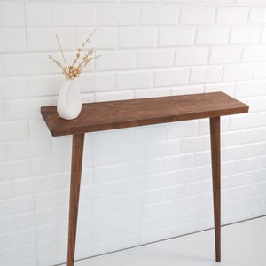 May include: A mid-century modern style console table with a dark brown wooden top and three slender, tapered legs. A white vase with dried flowers sits on the table. The table is against a white brick wall.