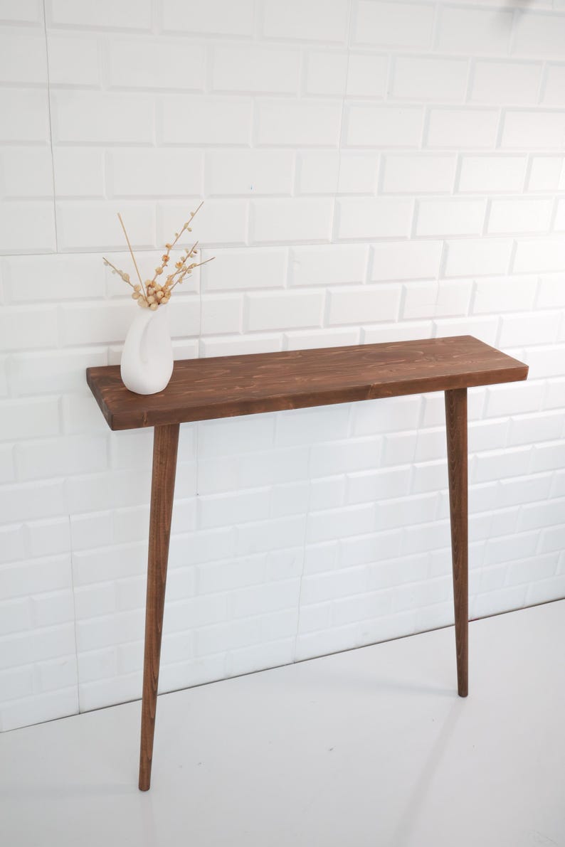 May include: A mid-century modern style console table with a dark brown wooden top and three slender, tapered legs. A white vase with dried flowers sits on the table. The table is against a white brick wall.