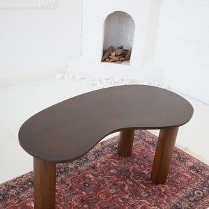 May include: A dark brown, kidney-shaped coffee table with four cylindrical wooden legs. The table sits on a patterned red and black rug, with a white fireplace in the background.