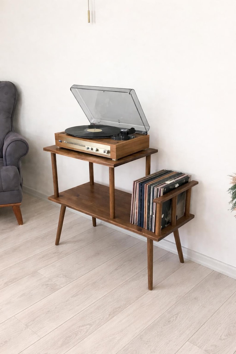 May include: A two-tiered wooden record player stand with tapered legs. The top tier holds a wooden turntable with a clear plastic dust cover. The lower tier features a built-in rack containing a collection of vinyl records in various sleeves.