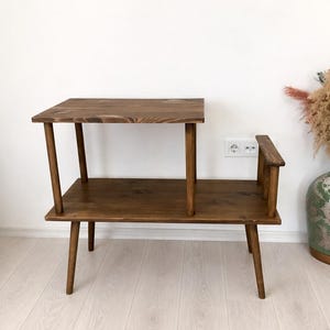 May include: A two-tiered wooden side table with a dark brown finish. The table features a rectangular lower shelf supported by four tapered legs, with a smaller rectangular upper shelf positioned on the left side, held up by two vertical wooden posts.