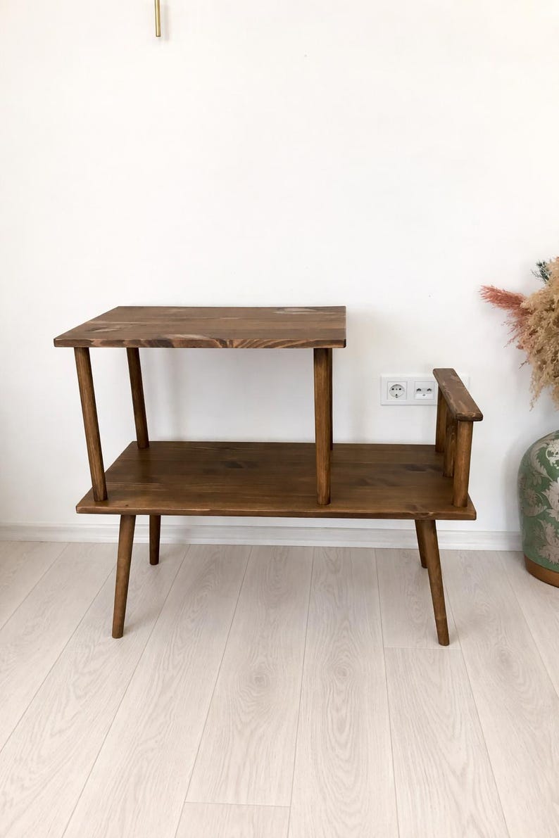 May include: A two-tiered wooden side table with a dark brown finish. The table features a rectangular lower shelf supported by four tapered legs, with a smaller rectangular upper shelf positioned on the left side, held up by two vertical wooden posts.