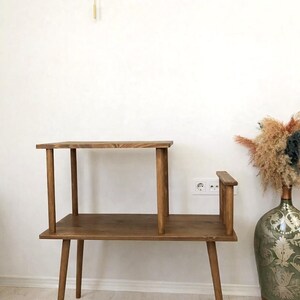 May include: A two-tiered wooden side table with a dark brown finish, featuring four tapered legs, a lower rectangular shelf, and a smaller upper shelf supported by two vertical posts, with a small vertical wooden accent piece on the right side.