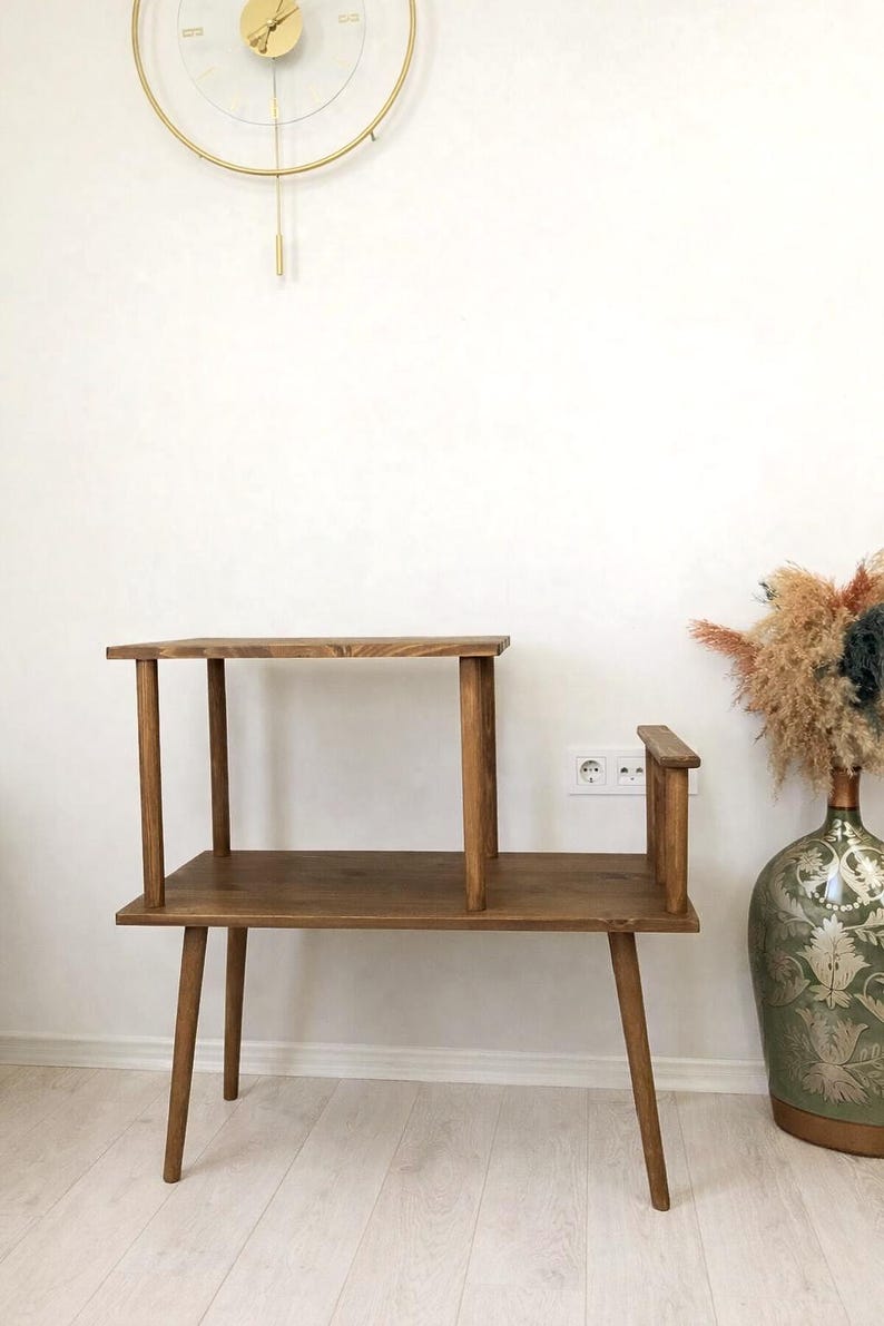 May include: A two-tiered wooden side table with a dark brown finish, featuring four tapered legs, a lower rectangular shelf, and a smaller upper shelf supported by two vertical posts, with a small vertical wooden accent piece on the right side.
