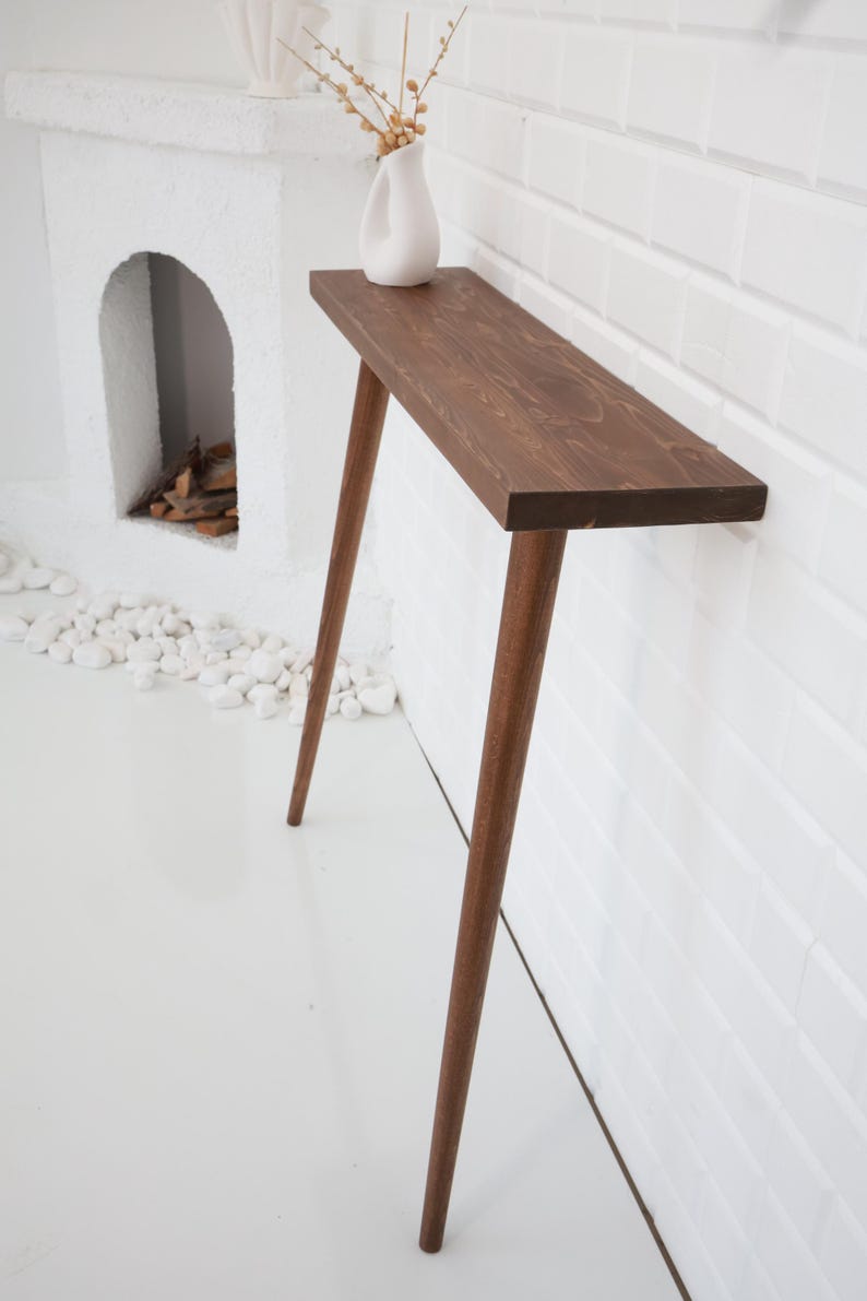 May include: A narrow, wooden console table with a dark brown finish. The table has a rectangular top and four slender, angled legs. A white vase with dried flowers sits on the table. The table is placed against a white brick wall and a white floor.