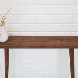 May include: A dark brown wooden console table with a rectangular top and four tapered legs. A white ceramic vase with a handle holds dried floral stems. The table is set against a white brick wall.