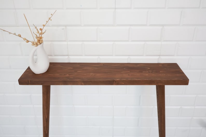 May include: A dark brown wooden console table with a rectangular top and four tapered legs. A white ceramic vase with a handle holds dried floral stems. The table is set against a white brick wall.