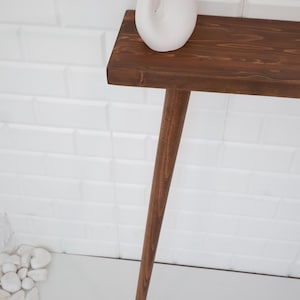 May include: A dark brown wooden console table with a single leg. A white ceramic vase with dried flowers sits on the table. The table is against a white brick wall.