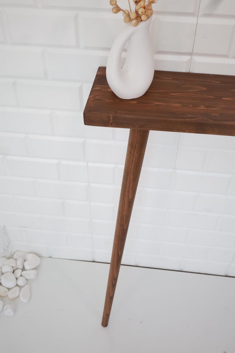 May include: A dark brown wooden console table with a single leg. A white ceramic vase with dried flowers sits on the table. The table is against a white brick wall.