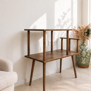 May include: A dark brown wooden tiered side table featuring a rectangular lower surface supported by four tapered legs and a smaller rectangular upper shelf held by four cylindrical posts, with an additional small side arm extending from the top.