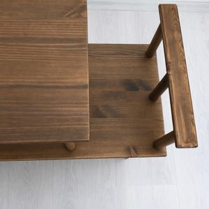 May include: A multi-tiered wooden stand featuring a dark brown stained finish with visible wood grain. The structure consists of a rectangular top surface, a lower shelf, and a vertical side handle supported by three cylindrical wooden posts.