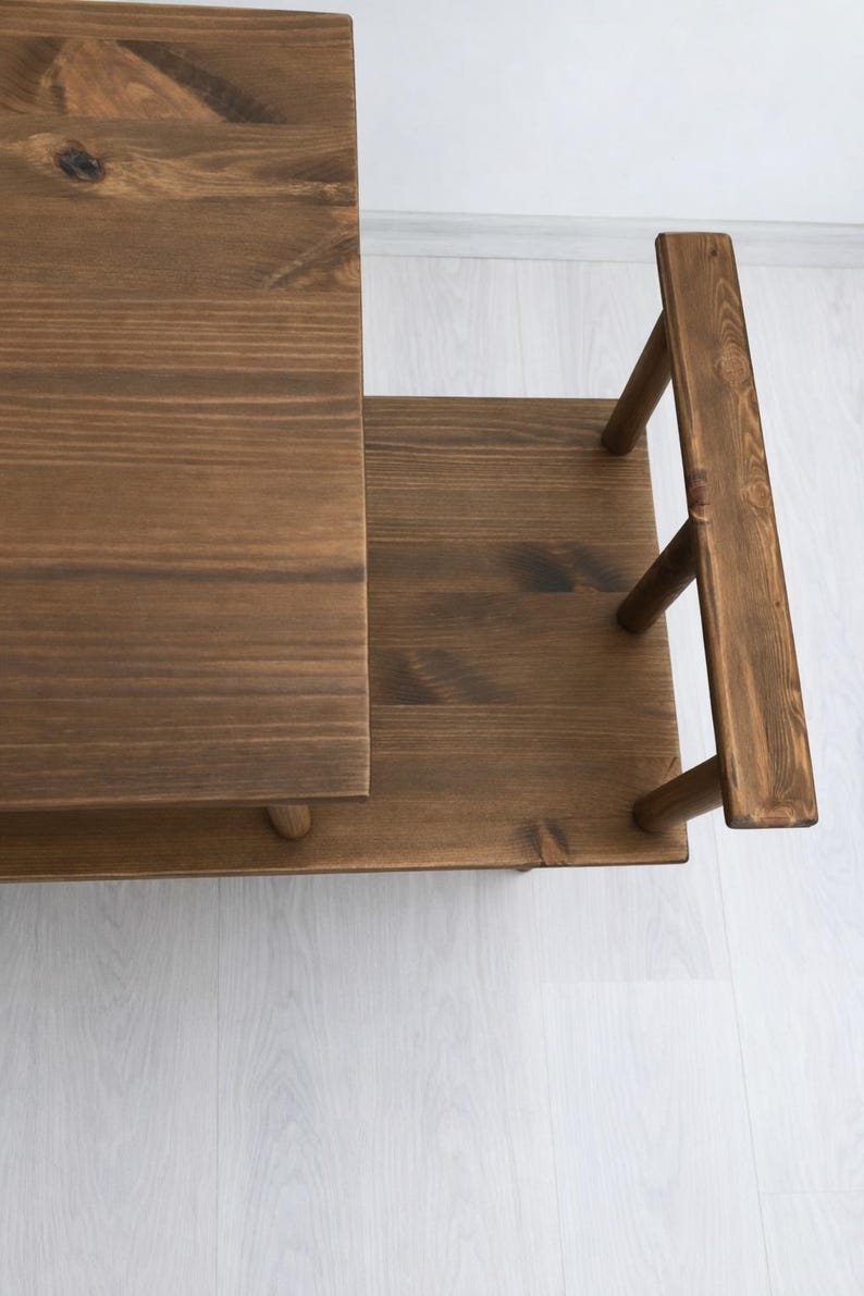 May include: A multi-tiered wooden stand featuring a dark brown stained finish with visible wood grain. The structure consists of a rectangular top surface, a lower shelf, and a vertical side handle supported by three cylindrical wooden posts.
