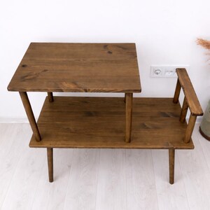 May include: A two-tiered wooden side table with a dark brown stained finish. The table features a rectangular lower shelf and a smaller, elevated rectangular upper surface supported by four wooden legs, with a small vertical wooden accent piece.
