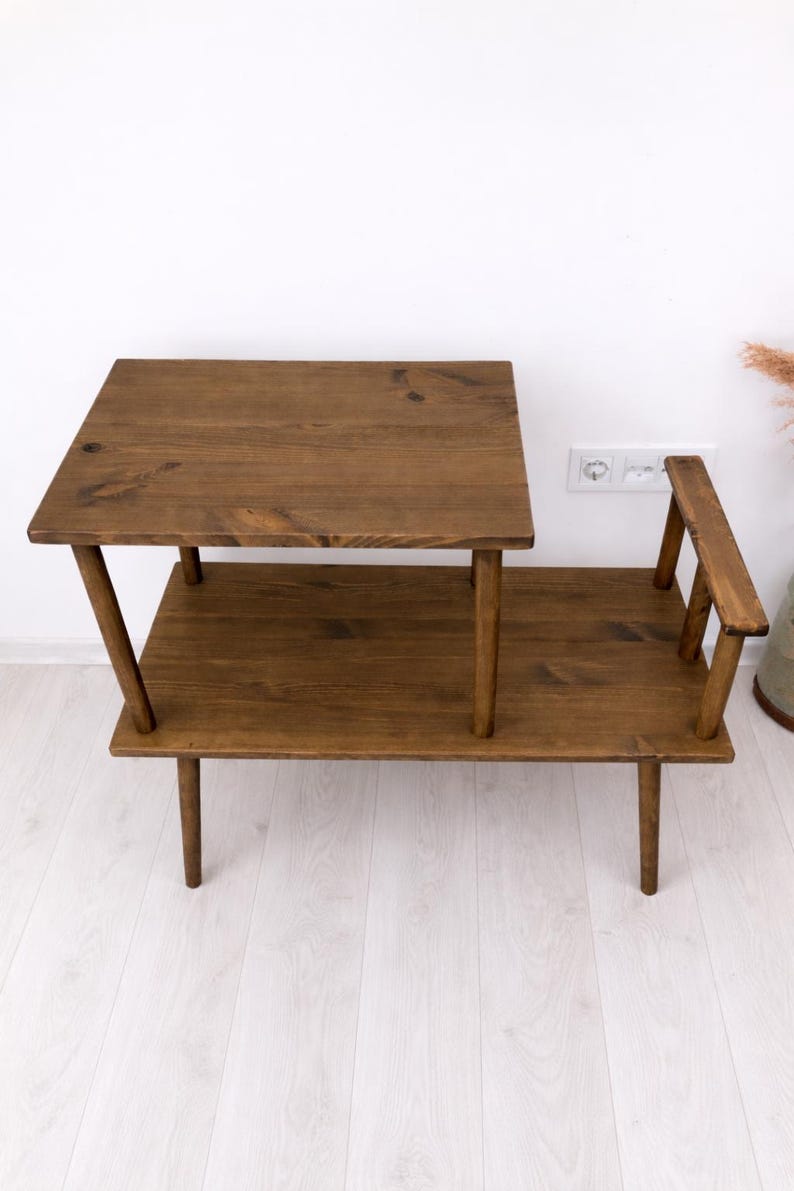 May include: A two-tiered wooden side table with a dark brown stained finish. The table features a rectangular lower shelf and a smaller, elevated rectangular upper surface supported by four wooden legs, with a small vertical wooden accent piece.