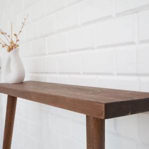 May include: A brown wooden console table with a rectangular top and four legs. A white vase with a unique shape holds dried branches with small, round, beige buds. The table is set against a white tiled wall.